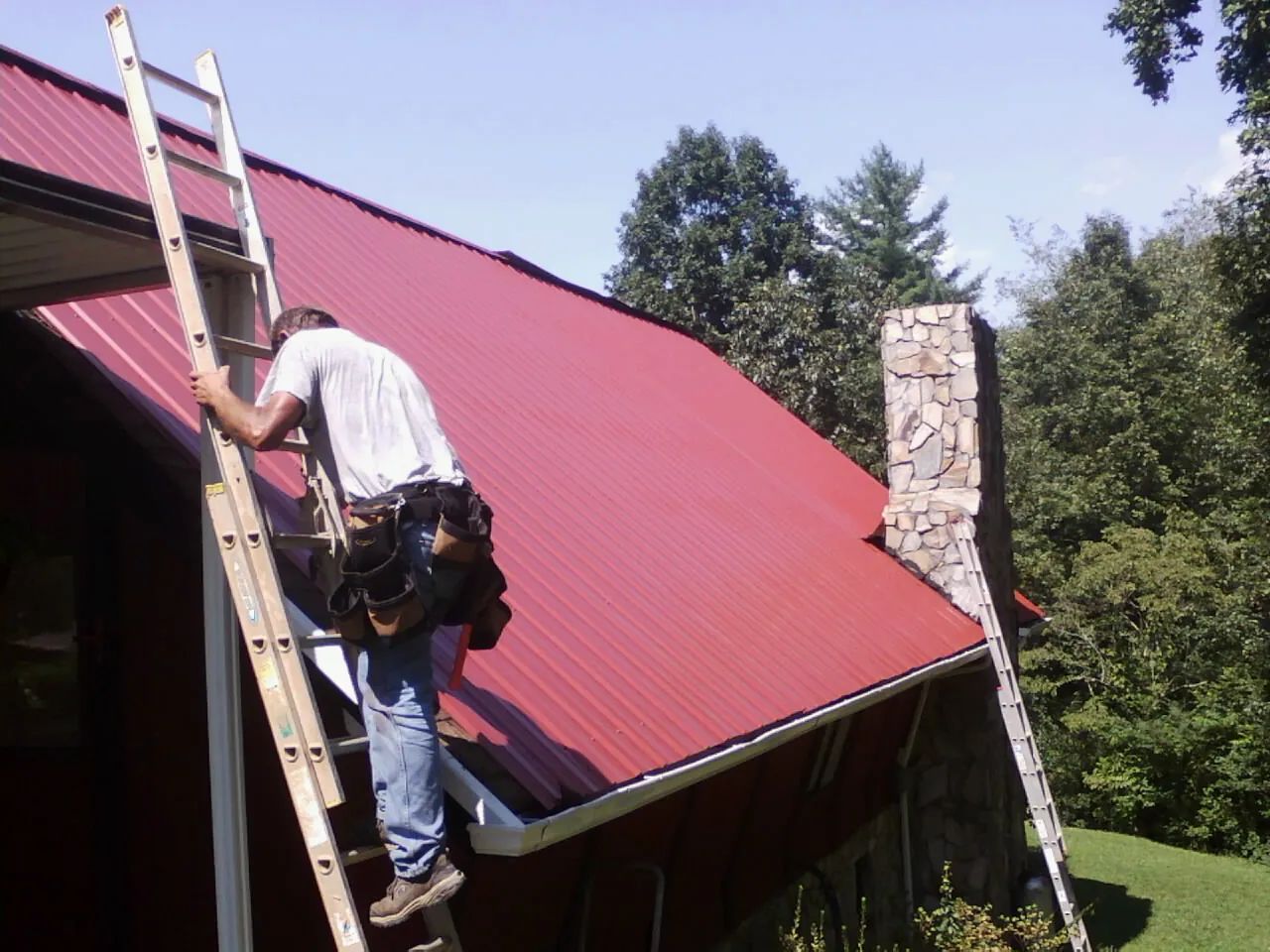 Roofer on a ladder beside a red metal roof and stone chimney — Soffit & Fascia Repair by Roofing Brothers in New Braintree